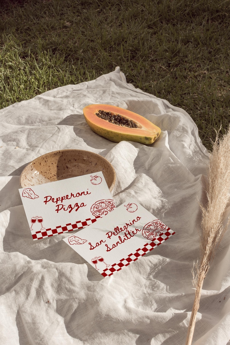 K&ouml;nnte beinhalten: Eine Picknick-Szene mit einem wei&szlig;en Tuch auf Gras. Eine halbierte Papaya, eine Keramikschale und zwei Karten mit rotem Text und Illustrationen sind zu sehen. Auf den Karten steht "Pepperoni Pizza" und "San Pellegrino Sanbitter".