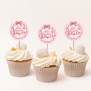 Pode incluir: Tr&ecirc;s cupcakes com cobertura branca, cada um coberto com um palito circular rosa que diz "She's Ready to Pop!" em letras vermelhas. Os cupcakes t&ecirc;m uma bola de coco branca e uma estrela dourada em cima.