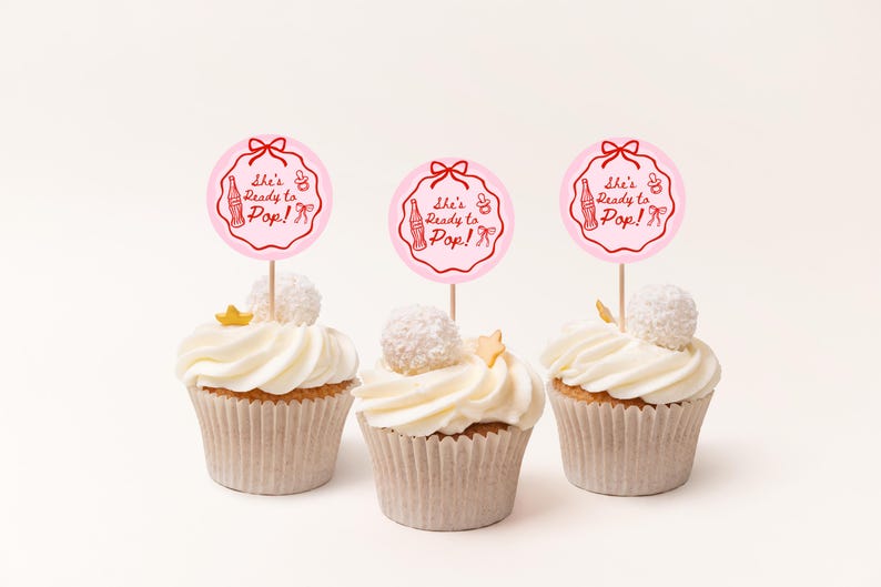 Pode incluir: Tr&ecirc;s cupcakes com cobertura branca, cada um coberto com um palito circular rosa que diz "She's Ready to Pop!" em letras vermelhas. Os cupcakes t&ecirc;m uma bola de coco branca e uma estrela dourada em cima.