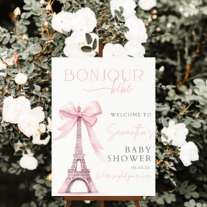 Puede incluir: Un cartel de bienvenida blanco con texto rosa y un lazo rosa en la parte superior de una Torre Eiffel rosa. El texto dice "Bonjour b&eacute;b&eacute;", "Bienvenido al Baby Shower de Samantha", y "06.03.24". El cartel tambi&eacute;n dice "Estamos encantados de que est&eacute;s aqu&iacute;".