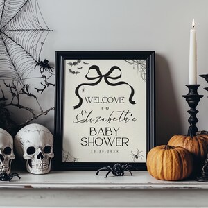 K&ouml;nnte beinhalten: Ein schwarzer Bilderrahmen mit dem Text "WELCOME to Elizabeth's BABY SHOWER" in schwarzer Schrift. Das Schild ist mit einer Schleife, Flederm&auml;usen und Spinnen verziert und von Halloween-Dekorationen wie K&uuml;rbissen und Totenk&ouml;pfen umgeben.