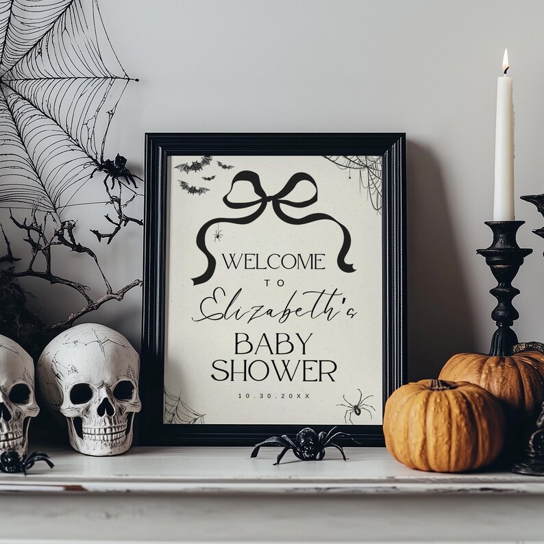 K&ouml;nnte beinhalten: Ein schwarzer Bilderrahmen mit dem Text "WELCOME to Elizabeth's BABY SHOWER" in schwarzer Schrift. Das Schild ist mit einer Schleife, Flederm&auml;usen und Spinnen verziert und von Halloween-Dekorationen wie K&uuml;rbissen und Totenk&ouml;pfen umgeben.