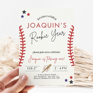 May include: Baseball-themed birthday party invitation with red stitching around the edges, a baseball bat and ball, and the text "Let's have a ball at Joaquin's Rookie Year. Please join us to celebrate Joaquin is turning one! Feb 27, 2-4 PM. At the Chester Fields, 123 Anystreet, Any City, CA. RSVP to Melissa 123.456.7891."