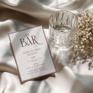 May include: A rectangular bar menu card with a dark red border and dark red text that reads The Bar, Signature Drinks, Wine, Beer, and Non Alcoholic, accompanied by a clear glass tumbler and a small bunch of dried white baby's breath flowers.
