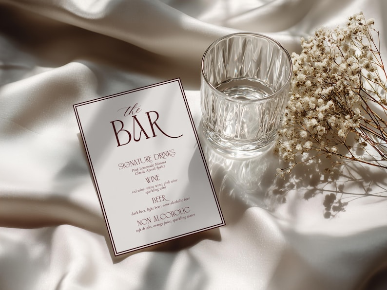 May include: A rectangular bar menu card with a dark red border and dark red text that reads The Bar, Signature Drinks, Wine, Beer, and Non Alcoholic, accompanied by a clear glass tumbler and a small bunch of dried white baby's breath flowers.