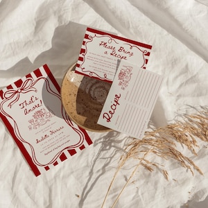 May include: Red and white striped invitation cards with the text "That's amore!" and "Please Bring a Recipe". The cards are on a white fabric background with a dried flower sprig.
