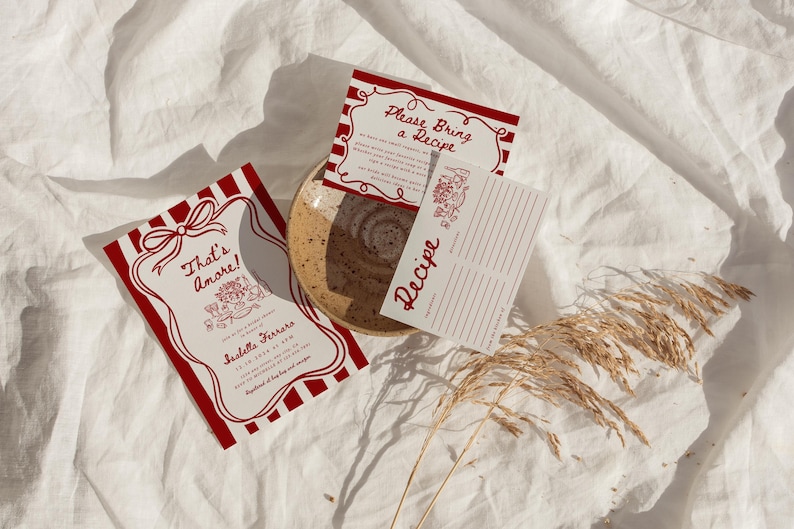 May include: Red and white striped invitation cards with the text "That's amore!" and "Please Bring a Recipe". The cards are on a white fabric background with a dried flower sprig.