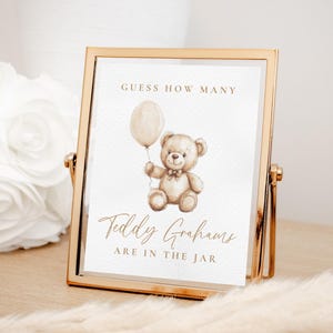 May include: A framed sign with a gold-colored metal frame. The sign features a watercolor illustration of a teddy bear holding a balloon. The text reads "Guess How Many Teddy Grahams Are In The Jar."
