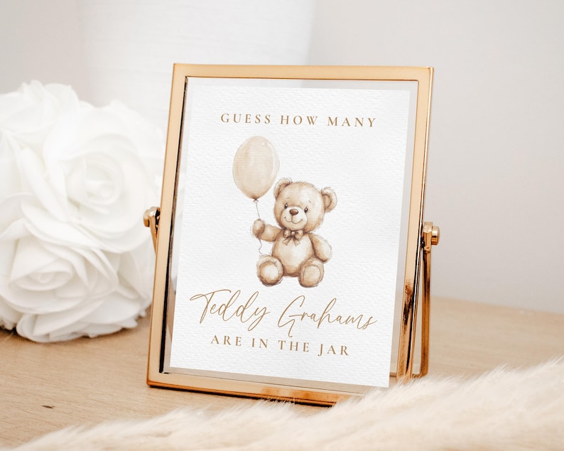 May include: A framed sign with a gold-colored metal frame. The sign features a watercolor illustration of a teddy bear holding a balloon. The text reads "Guess How Many Teddy Grahams Are In The Jar."