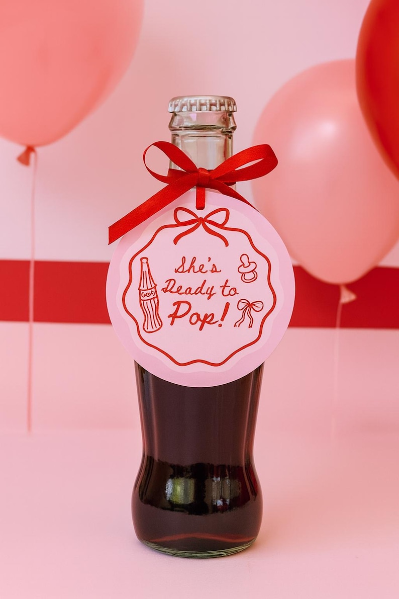 Pode incluir: Uma garrafa de vidro cheia de um l&iacute;quido escuro, adornada com uma etiqueta rosa e uma fita vermelha. A etiqueta diz "She's Ready to Pop!" com uma ilustra&ccedil;&atilde;o de biber&atilde;o e la&ccedil;o. Bal&otilde;es cor-de-rosa no fundo.