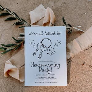 K&ouml;nnte beinhalten: Eine wei&szlig;e Einladung zu einer Einweihungsparty mit einer schwarz-wei&szlig;en Illustration von Schl&uuml;sseln und einem Haus. Der Text lautet "We're all settled in!" und "Housewarming Party!" Das Datum und die Uhrzeit der Party sind der 06. Oktober 2024 um 15:00 Uhr. Die Adresse und die RSVP-Informationen sind ebenfalls enthalten. Die Einladung gibt an, dass das Paar bei Target und Amazon registriert ist.