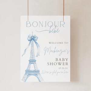 May include: A blue and white baby shower welcome sign with a watercolour illustration of the Eiffel Tower and a bow. The sign says "Bonjour Bibi" and "Welcome to Mackenzie's Baby Shower 07.20.24".