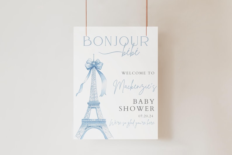 May include: A blue and white baby shower welcome sign with a watercolour illustration of the Eiffel Tower and a bow. The sign says "Bonjour Bibi" and "Welcome to Mackenzie's Baby Shower 07.20.24".