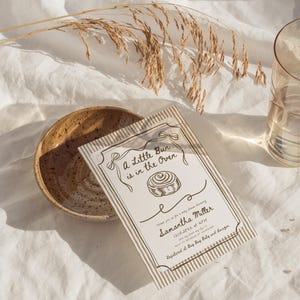 May include: A baby shower invitation with the text "A Little Bun is in the Oven" and the name Samantha Miller. The invitation is on a white background with a striped border and a drawing of a cinnamon roll. A ceramic bowl and a glass of water are also in the image.