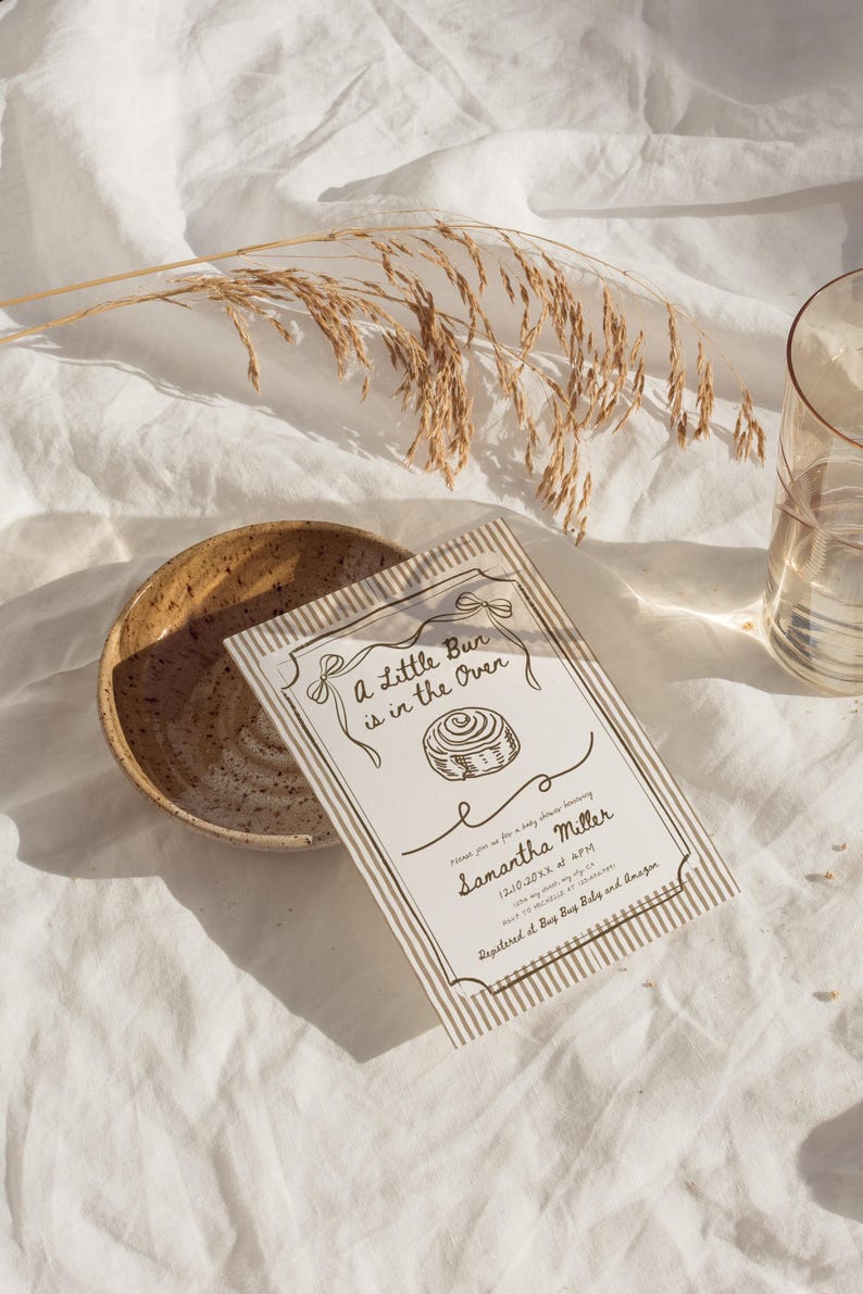 May include: A baby shower invitation with the text "A Little Bun is in the Oven" and the name Samantha Miller. The invitation is on a white background with a striped border and a drawing of a cinnamon roll. A ceramic bowl and a glass of water are also in the image.
