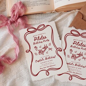 May include: Two cream-colored Pilates birthday party invitations with a burgundy border and text. The invitations feature illustrations of Pilates equipment and text that reads "Crystal's Birthday!" and "Dress Code: Cute Activewear."