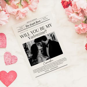 May include: A Valentine's Day themed image featuring a newspaper-style print with the headline "WILL YOU BE MY Valentine?" and a black and white photo of a couple kissing. Pink heart cutouts and flowers surround the print.
