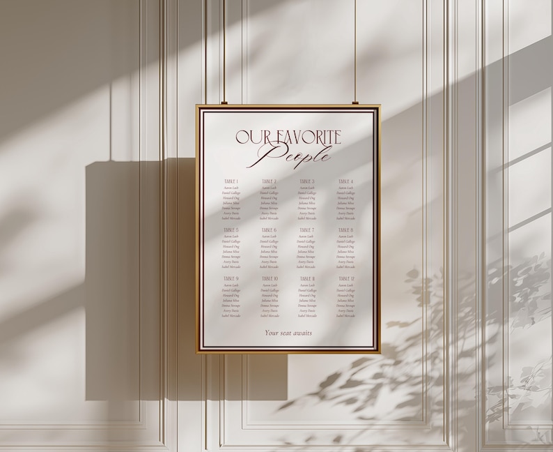 May include: A rectangular wedding seating chart sign with a gold frame, featuring the text "OUR FAVORITE PEOPLE" at the top and "Your seat awaits" at the bottom. The white sign displays twelve tables with lists of names in dark red serif font.