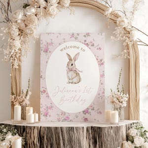 May include: A pink floral welcome sign with a white oval featuring a brown bunny with a pink bow. The text on the sign reads "welcome to Julianna's 1st Birthday".