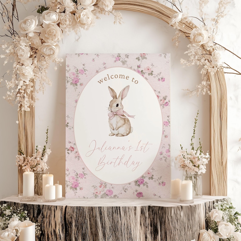 May include: A pink floral welcome sign with a white oval featuring a brown bunny with a pink bow. The text on the sign reads "welcome to Julianna's 1st Birthday".