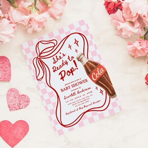 Puede incluir: Una invitaci&oacute;n a un baby shower con un dise&ntilde;o retro. La invitaci&oacute;n presenta un fondo a cuadros rojos y blancos, un contorno rojo y el texto "She's Ready to Pop!". Se incluye una ilustraci&oacute;n de una botella de cola. Decoraciones en forma de coraz&oacute;n rosa y flores rodean la invitaci&oacute;n.