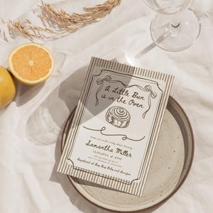 May include: Baby shower invitation with the text "A Little Bun is in the Oven" and the name "Samantha Miller". The invitation is on a plate with a glass of water and a lemon half.
