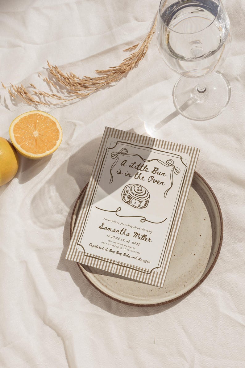 May include: Baby shower invitation with the text "A Little Bun is in the Oven" and the name "Samantha Miller". The invitation is on a plate with a glass of water and a lemon half.
