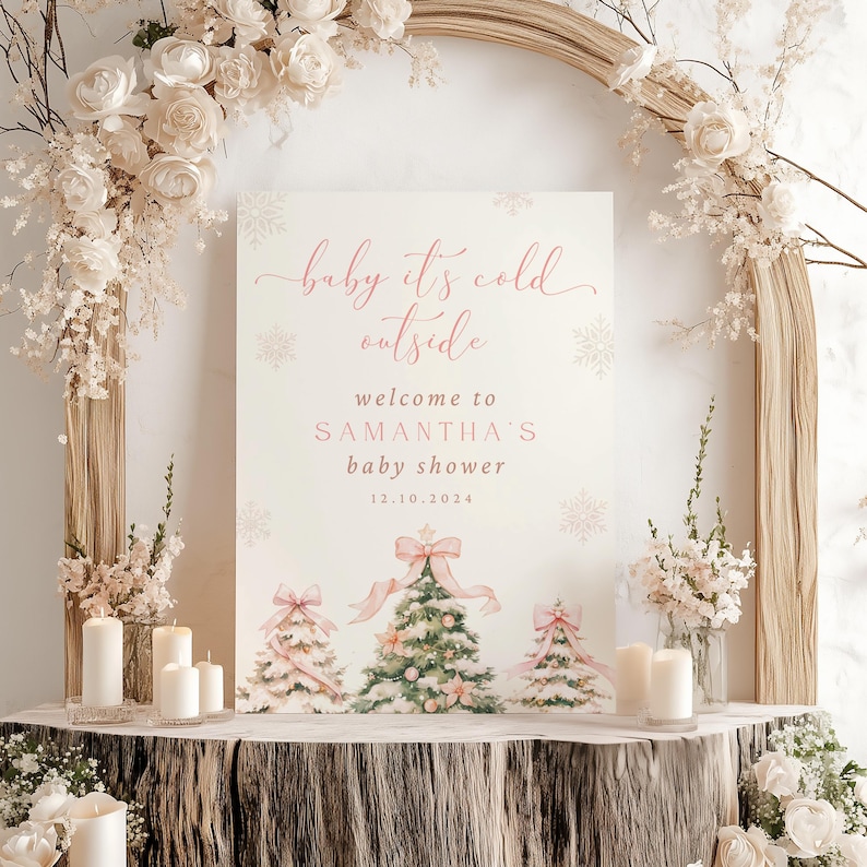 May include: A white sign with pink text that reads "baby it's cold outside" and "welcome to Samantha's baby shower 12.10.2024". The sign features a watercolor illustration of three Christmas trees with pink and green decorations.