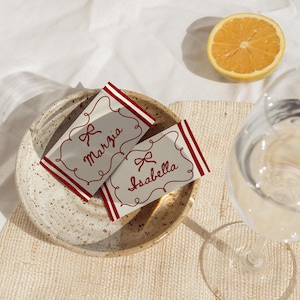Pu&ograve; includere: Due segnaposto con dettagli rossi e i nomi "Marzia" e "Isabella" sono su un piatto maculato. Mezzo limone e un bicchiere d'acqua sono anche nell'immagine. Le carte hanno un design a fiocco.