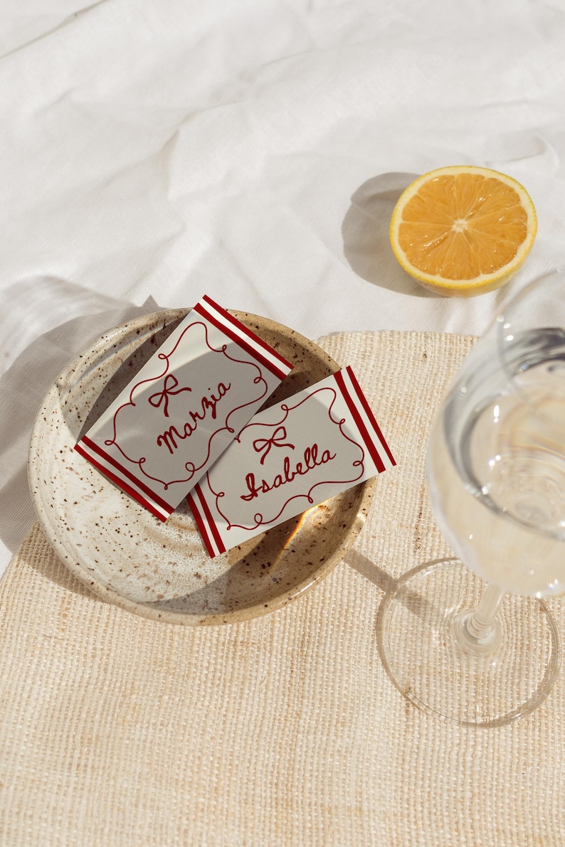 Pu&ograve; includere: Due segnaposto con dettagli rossi e i nomi "Marzia" e "Isabella" sono su un piatto maculato. Mezzo limone e un bicchiere d'acqua sono anche nell'immagine. Le carte hanno un design a fiocco.