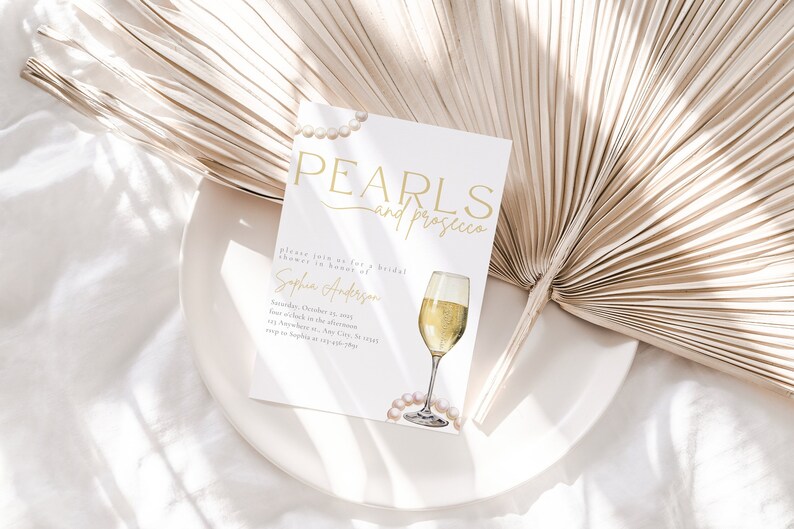 May include: A white bridal shower invitation with gold text that reads "Pearls and Prosecco". The invitation features a watercolor illustration of a glass of champagne and a string of pearls.