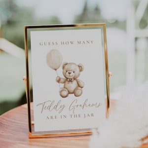 May include: A framed sign with gold trim, featuring a watercolor illustration of a teddy bear holding a balloon. The sign reads "Guess How Many" and "Teddy Grahams are in the jar."