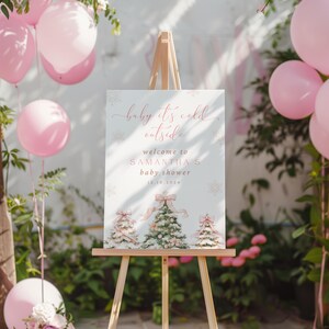 May include: A white sign with pink text that reads "baby it's cold outside, welcome to Samantha's baby shower 12.10.2014". The sign is decorated with snowflakes and three watercolor illustrations of Christmas trees with pink bows.