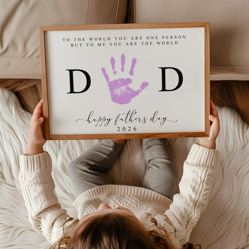 May include: A framed print with the text "TO THE WORLD YOU ARE ONE PERSON BUT TO ME YOU ARE THE WORLD" and "happy fathers day 2026". The print features the word "DAD" with a purple handprint in the center. The frame is made of wood.
