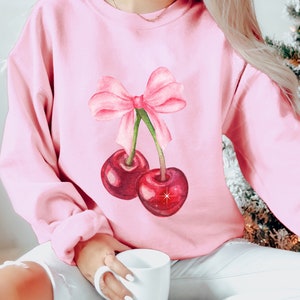May include: A pink sweatshirt with a watercolor illustration of two cherries with a pink bow on top.