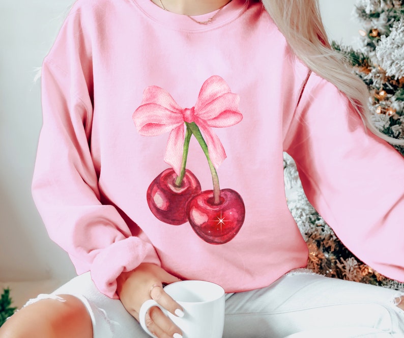 May include: A pink sweatshirt with a watercolor illustration of two cherries with a pink bow on top.