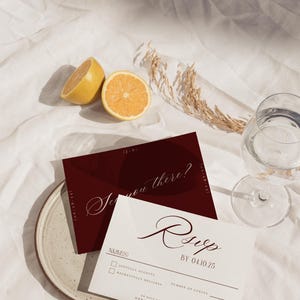 May include: A styled photograph featuring wedding stationery. A burgundy invitation with the text "See you there?" and a white RSVP card with the text "RSVP BY 04.10.25" are displayed. The scene includes a halved lemon, a glass of water, and decorative elements.