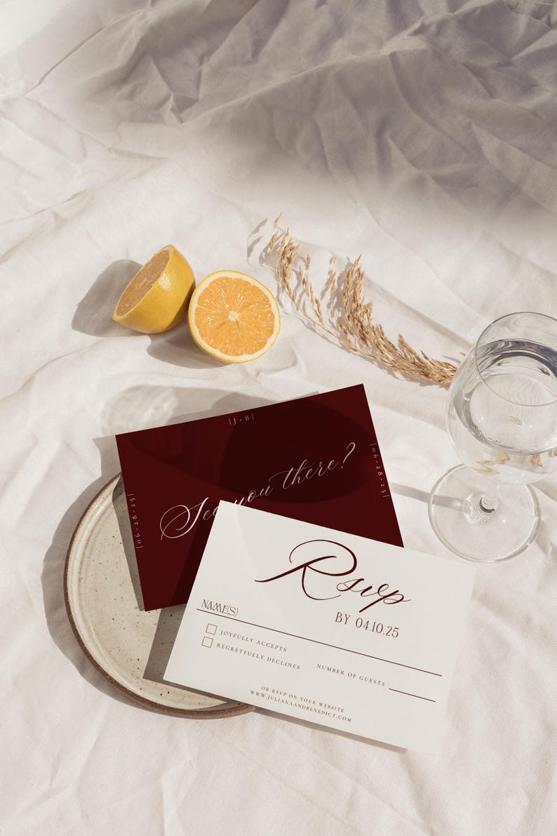 May include: A styled photograph featuring wedding stationery. A burgundy invitation with the text "See you there?" and a white RSVP card with the text "RSVP BY 04.10.25" are displayed. The scene includes a halved lemon, a glass of water, and decorative elements.