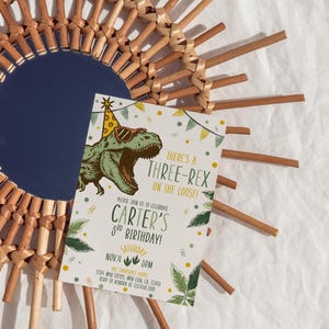 May include: A birthday invitation featuring a cartoon dinosaur wearing sunglasses and a party hat. The invitation reads "There's a Three-Rex on the Loose!" and announces Carter's 3rd birthday on November 4th at 3 PM. The invitation is set against a woven sunburst mirror.