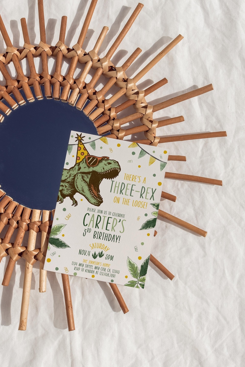 May include: A birthday invitation featuring a cartoon dinosaur wearing sunglasses and a party hat. The invitation reads "There's a Three-Rex on the Loose!" and announces Carter's 3rd birthday on November 4th at 3 PM. The invitation is set against a woven sunburst mirror.