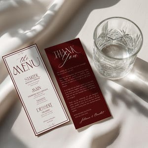 May include: A vertical menu card with cream paper and dark red borders, listing courses like Buffalo Caprese and Tiramisu, alongside a matching dark red thank you card with white text, positioned next to a clear, textured glass tumbler.