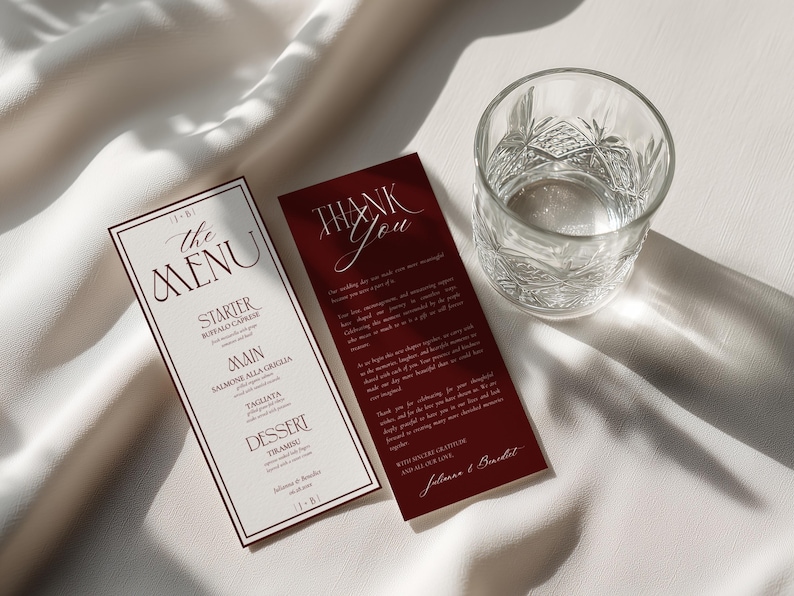 May include: A vertical menu card with cream paper and dark red borders, listing courses like Buffalo Caprese and Tiramisu, alongside a matching dark red thank you card with white text, positioned next to a clear, textured glass tumbler.