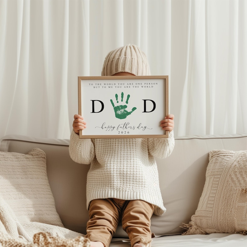 May include: A framed print with the text "TO THE WORLD YOU ARE ONE PERSON BUT TO ME YOU ARE THE WORLD" and "D D happy fathers day 2026". The print features a green handprint. The frame is a light brown color.