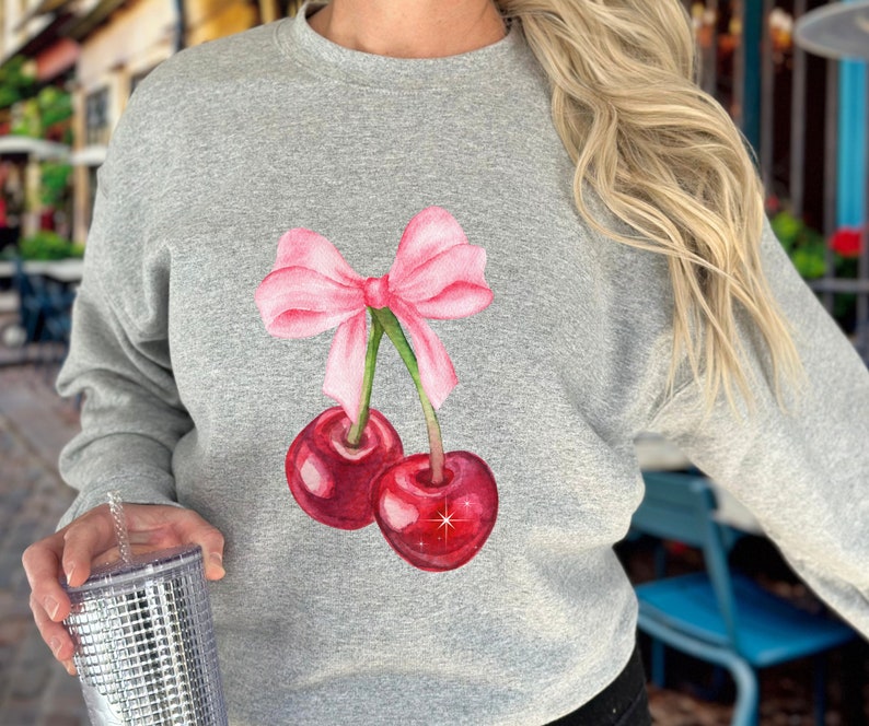 May include: A gray sweatshirt with a pink bow and two red cherries. The cherries have green stems and leaves.
