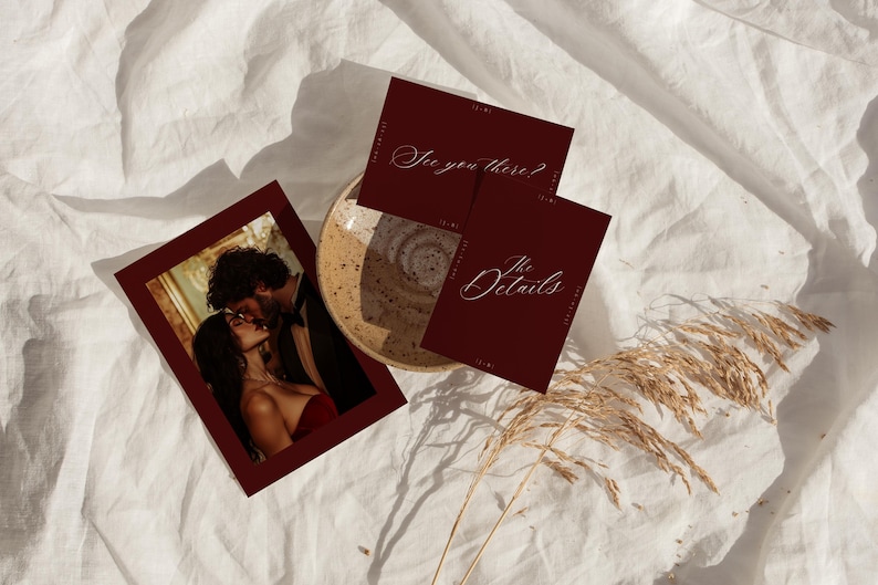 May include: A burgundy invitation suite with a photo of a couple kissing, and two cards with the phrases "See you there?" and "The Details" in elegant script. The items are arranged on a white, textured surface.