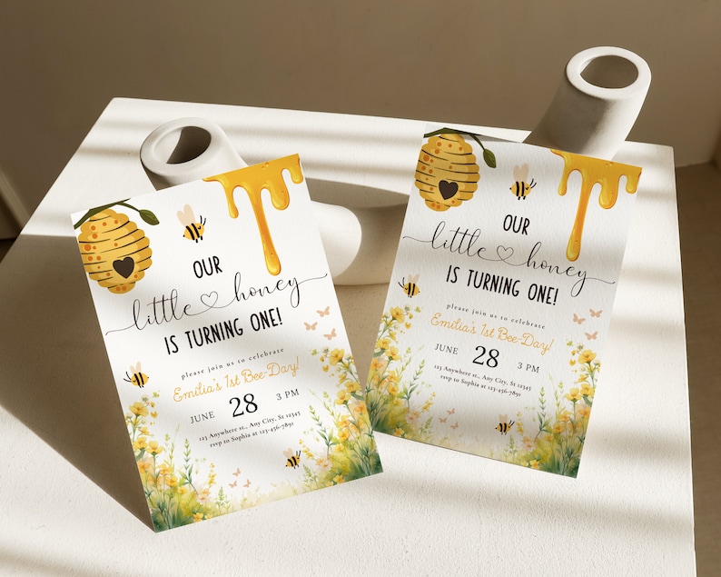 May include: Two yellow and white bee-themed first birthday party invitations with a honeycomb design, dripping honey, and a floral background. The text on the invitations reads "Our little honey is turning one!" and "Emilia's 1st Bee-Day!" with the date and time of the party.