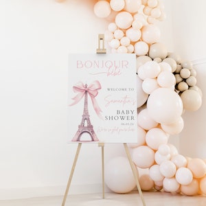 Puede incluir: Un cartel de bienvenida para una baby shower rosa y blanco con una ilustraci&oacute;n de acuarela de la Torre Eiffel y un lazo rosa. El cartel dice "Bonjour bebe, Bienvenida a la baby shower de Samantha, 06.03.24, Estamos encantados de que est&eacute;s aqu&iacute;."