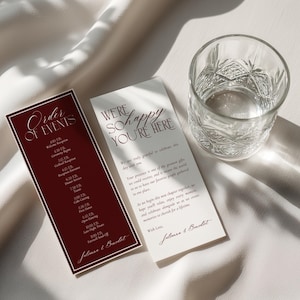May include: Two rectangular wedding stationery cards with dark red and cream backgrounds. One card features the text Order of Events and the other We're So Happy You're Here, both signed by Julianna & Benedict, positioned next to a clear glass tumbler.