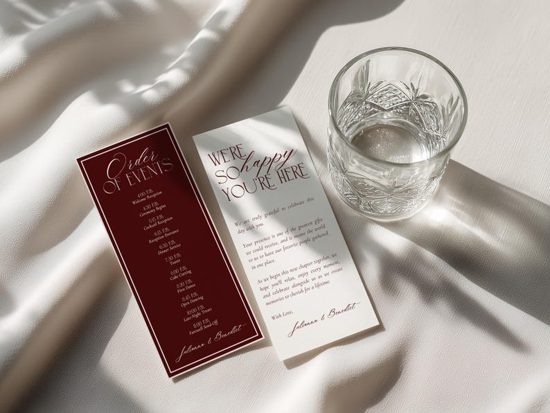 May include: Two rectangular wedding stationery cards with dark red and cream backgrounds. One card features the text Order of Events and the other We're So Happy You're Here, both signed by Julianna & Benedict, positioned next to a clear glass tumbler.