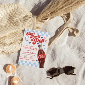 Puede incluir: Una invitaci&oacute;n a un baby shower con el texto "She's Ready to Pop" y una ilustraci&oacute;n de una botella de cola. La invitaci&oacute;n est&aacute; sobre un fondo a cuadros azul y blanco. Tambi&eacute;n se muestran gafas de sol, una bolsa de red, hierba seca y rodajas de naranja.